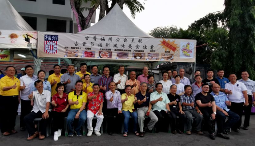 众人合影于古晋福州公会摊位前，前排左起为古晋福州公会财政陈其富、妇女组主任刘碧云、首长署政治秘书陈开、华总执行顾问黄张金发、古晋福州公会会长拿督余志豪、砂地方政府与房屋部长拿督沈桂贤、中国驻古晋副总领事张阳、古晋福州公会署理会长王长福、程洪领事、副会长李廷闻、青年团团长黄耀明和砂华青团总团长林学定。