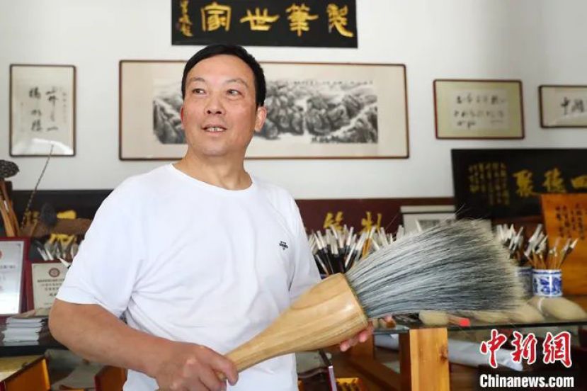 周鹏程手中拿着一支巨型毛笔，身后的牌匾上写着“制笔世家”四个字。